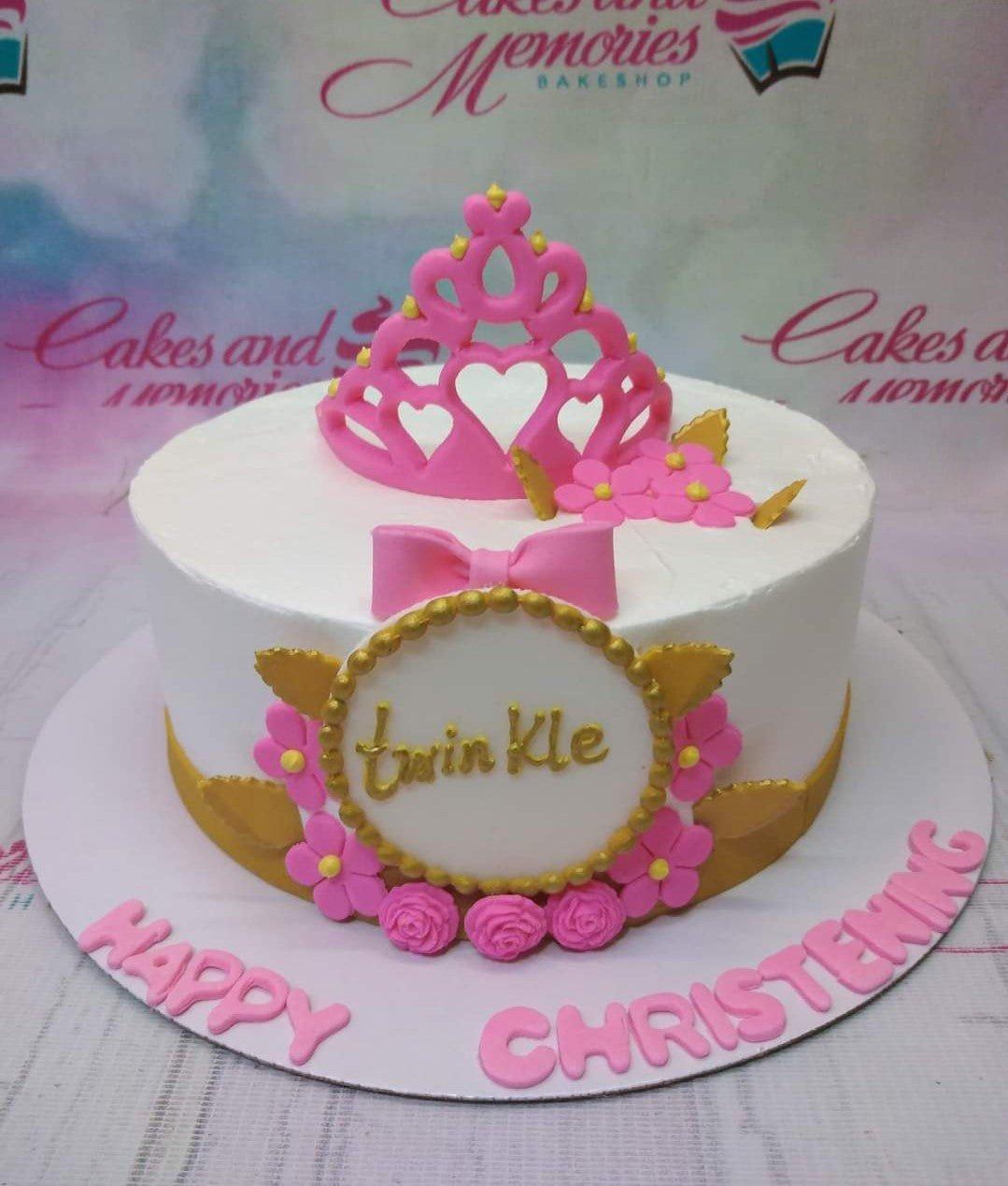 White round christening cake with a pink fondant crown, pink flowers, and gold leaf accents featuring the name Twinkle.