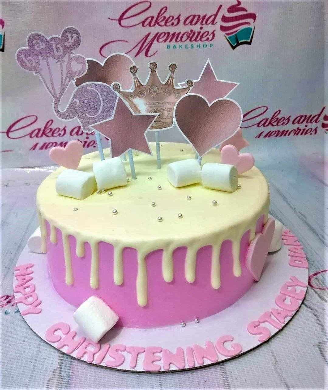 Pink and yellow christening cake with a crown, elephant, and heart toppers, decorated with marshmallows and silver dragees.