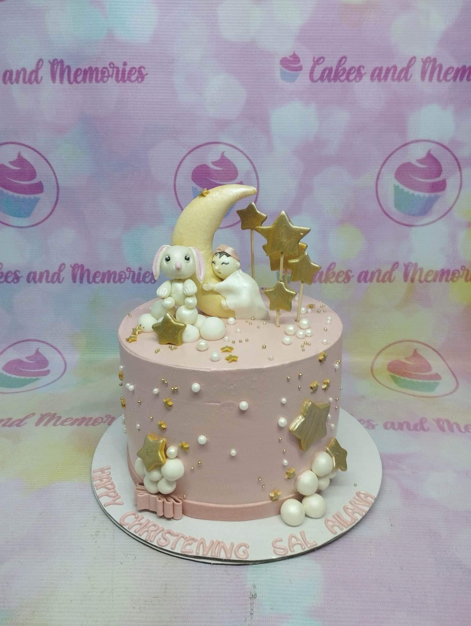 Pink christening cake with a fondant moon, sleeping baby, bunny, and gold stars.