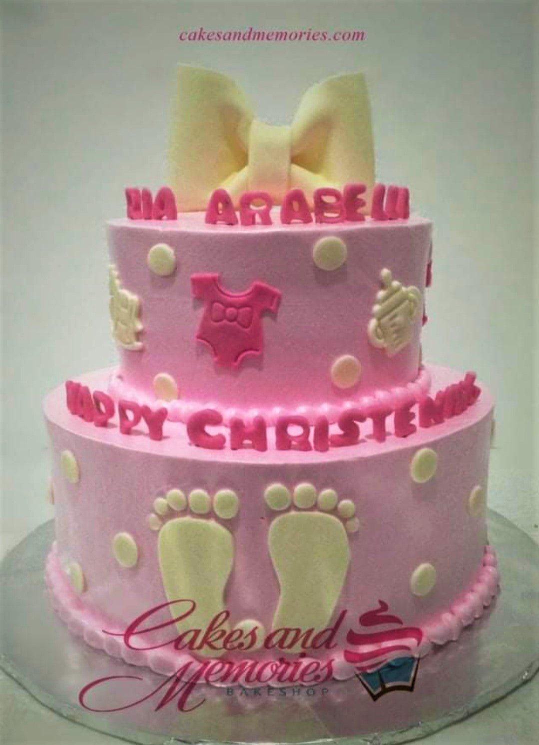 Pink 2-tier christening cake with a large yellow fondant bow, baby-themed decorations like footprints and onesies, and pink gumpaste letters.