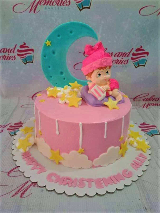 Pink christening cake with a large teal moon, a baby girl figurine, and yellow stars on a white board with pink lettering.
