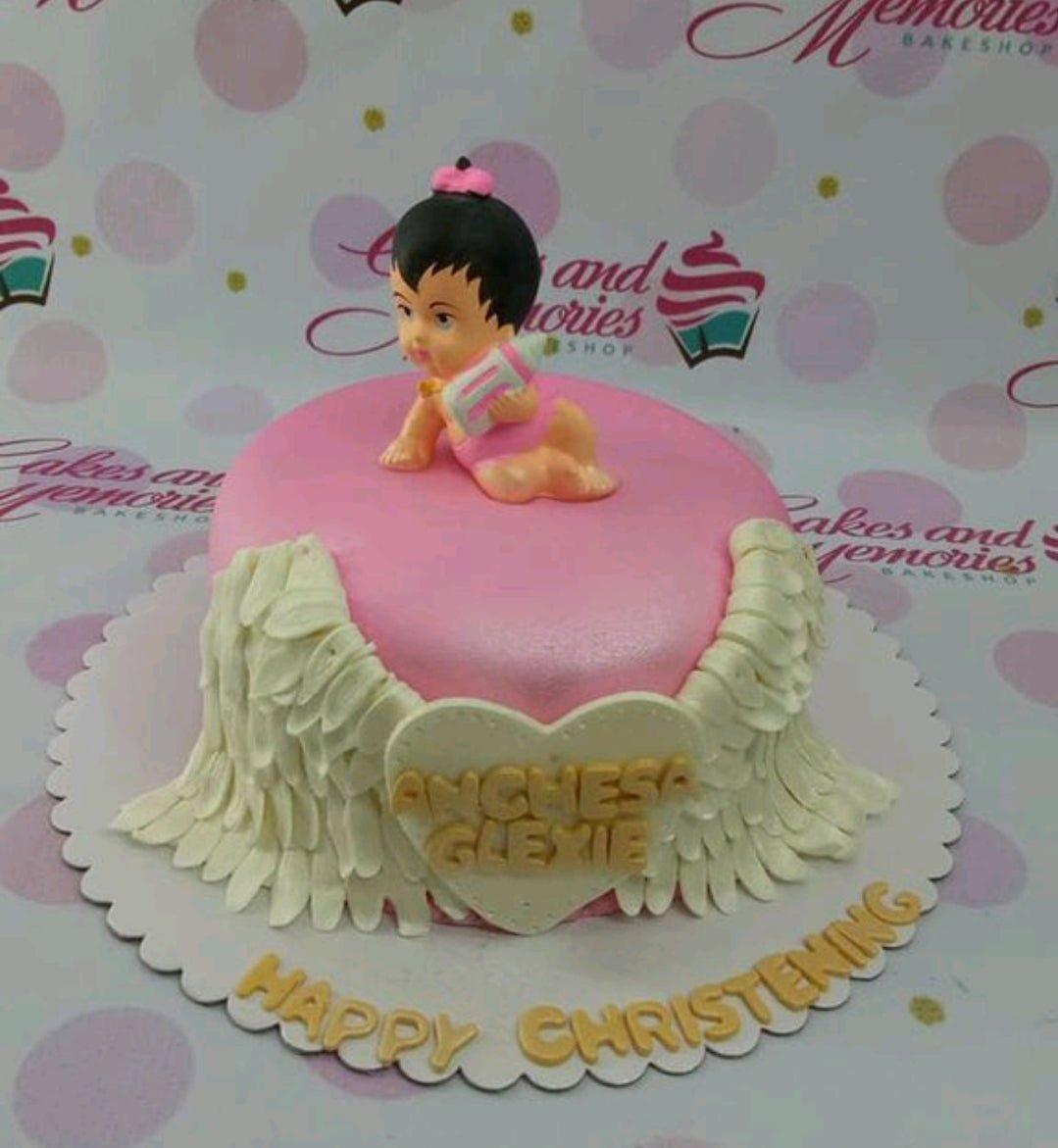 Pink fondant christening cake with a 3D baby girl topper, white angel wings, and a heart-shaped name plaque.