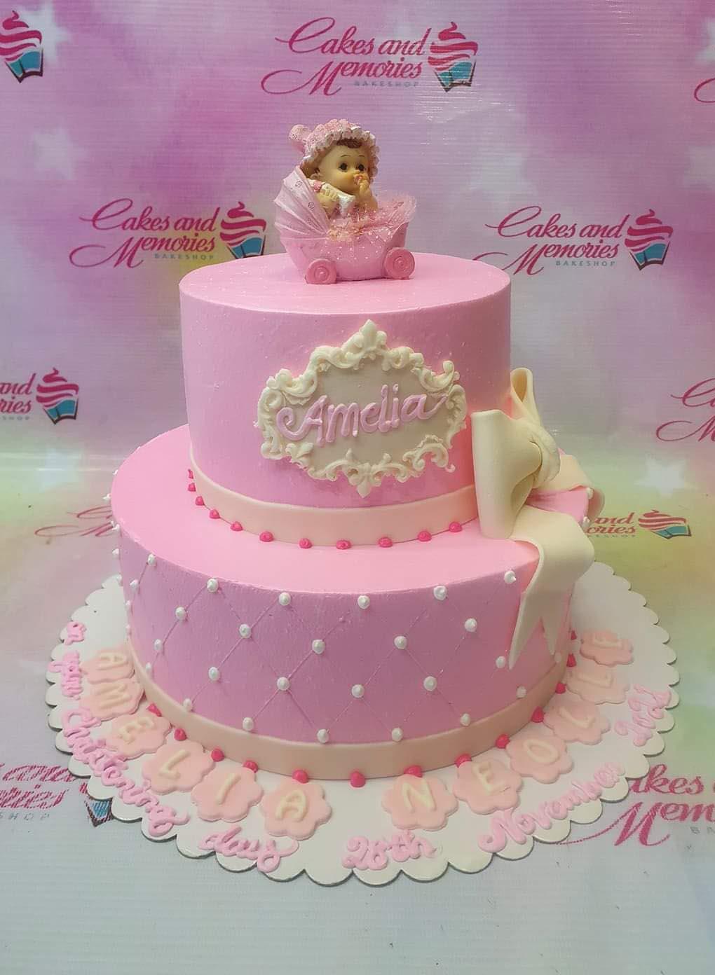Pink 2-tier baby christening cake with a fondant baby in a stroller topper, white bow, and quilted pattern with white pearls.