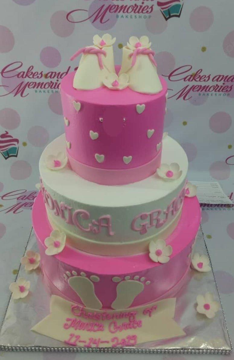 Pink and white 3-tier christening cake with fondant baby shoes, flowers, and baby footprints.