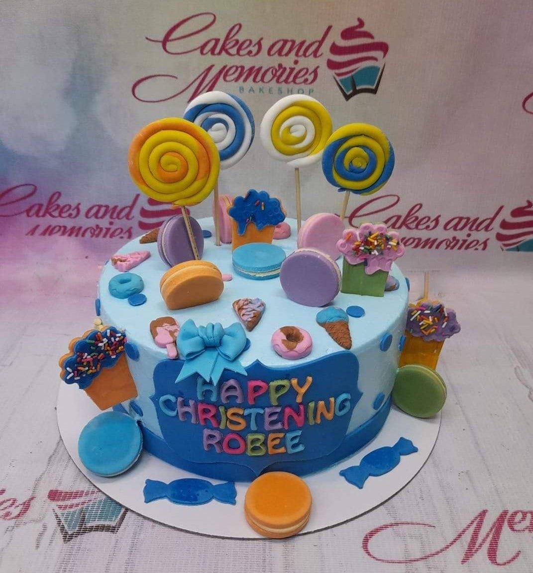 Light blue christening cake for Robee featuring colorful fondant lollipops, macarons, donuts, and ice cream cones with a blue bow.