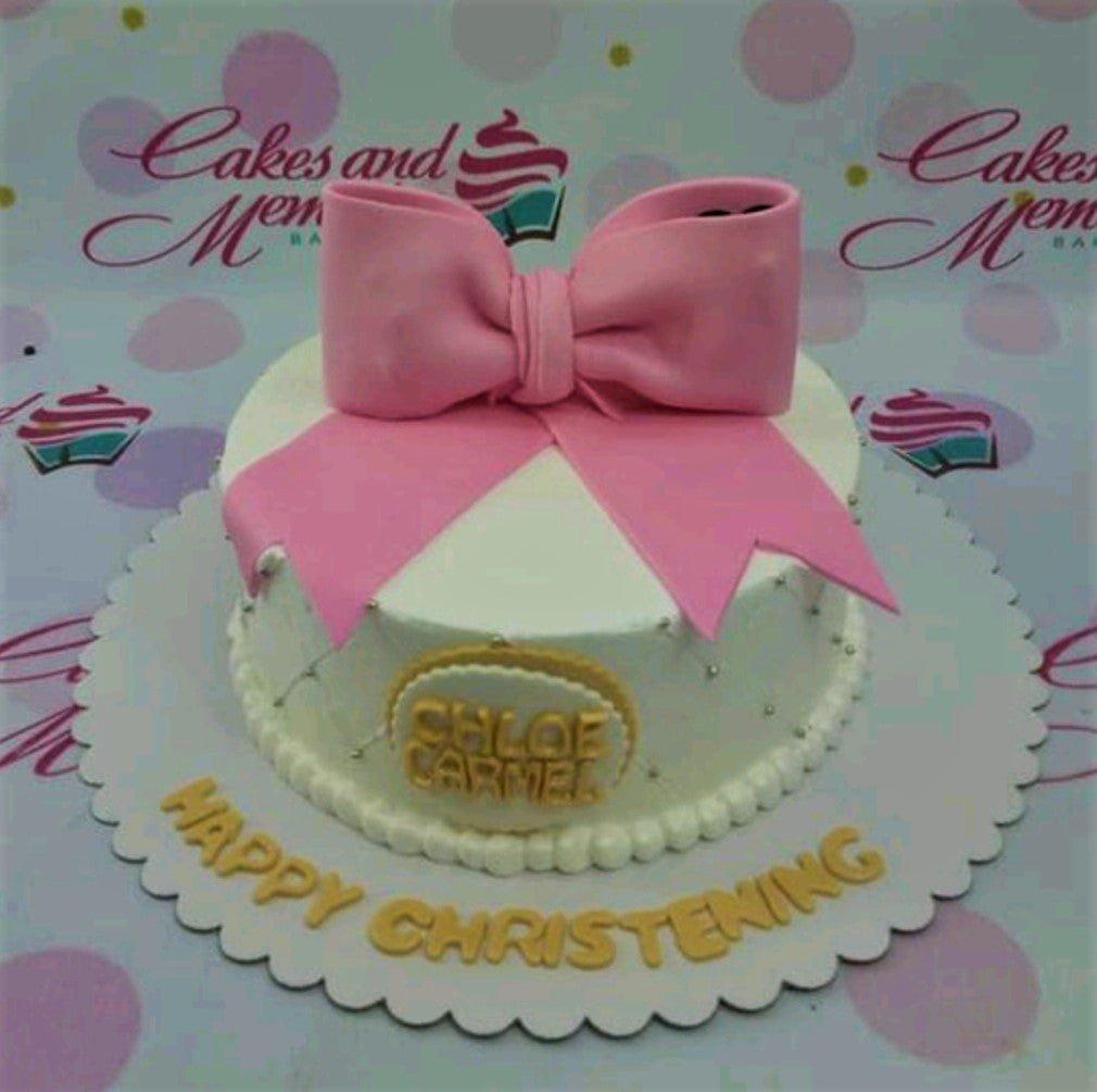 White round cake with a large pink fondant bow on top and gold gumpaste letters on the base board.