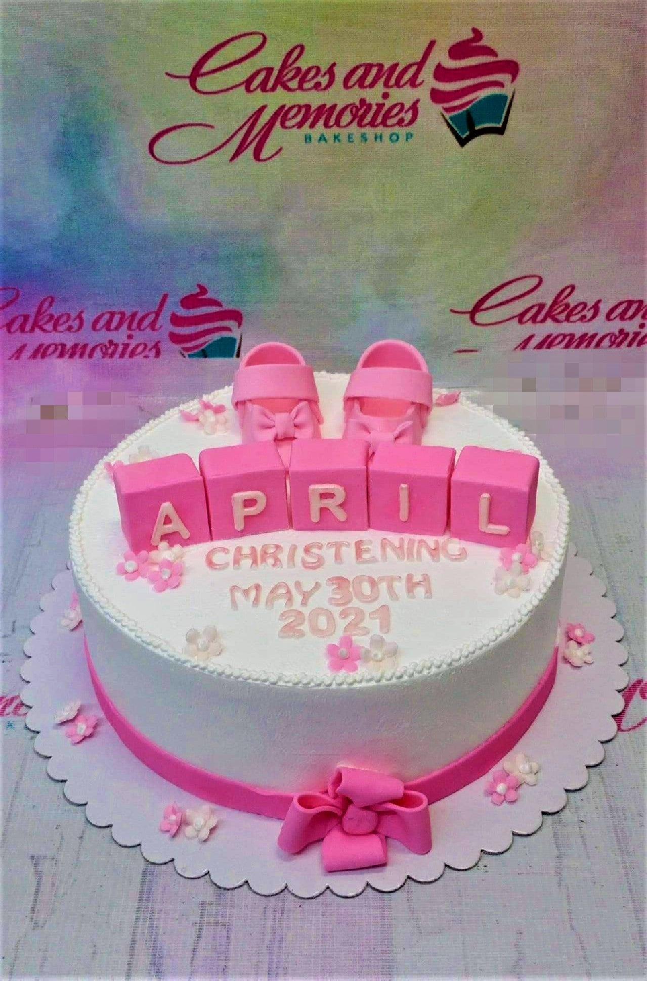 White 1-tier christening cake with pink fondant baby shoes, alphabet blocks spelling APRIL, and small pink and white flowers.