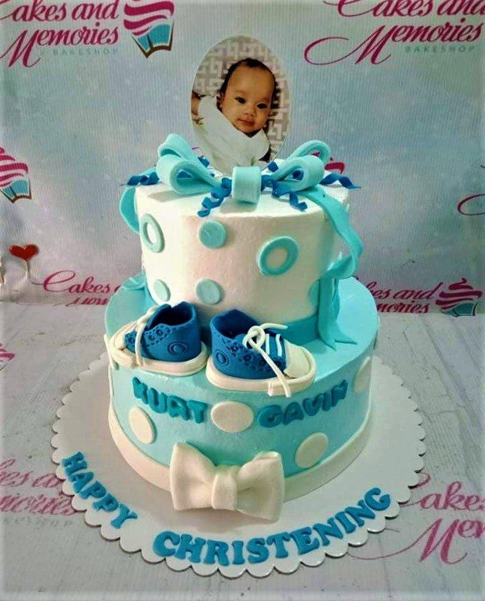 Two-tier light blue and white christening cake for a baby boy featuring a photo topper, fondant baby shoes, and white bows.