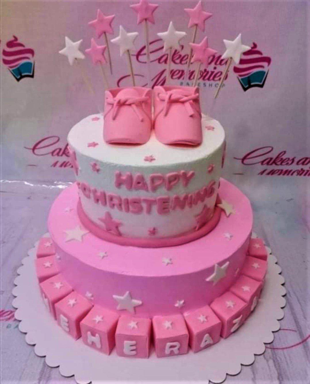Pink and white two-tier christening cake with pink baby booties, stars on sticks, and pink letter blocks for a baby girl.