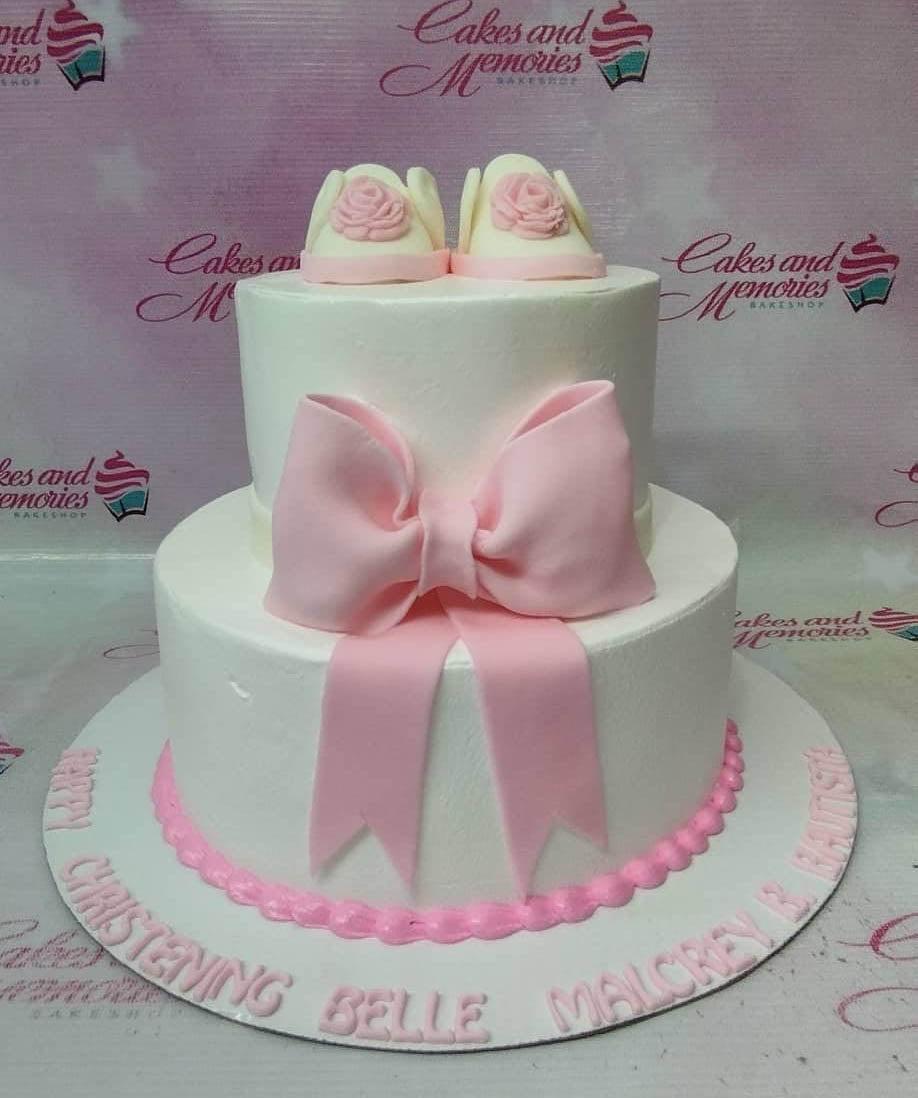 Two-tier white fondant christening cake with a large pink bow and edible pink baby shoes on top.