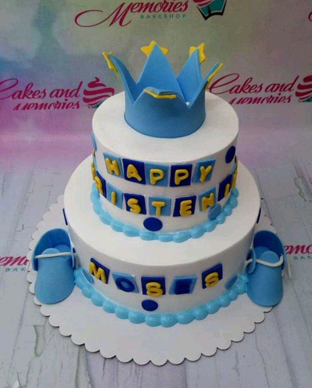 Two-tier white fondant cake with blue crown topper, blue baby shoes, and yellow gumpaste letters for a christening celebration.