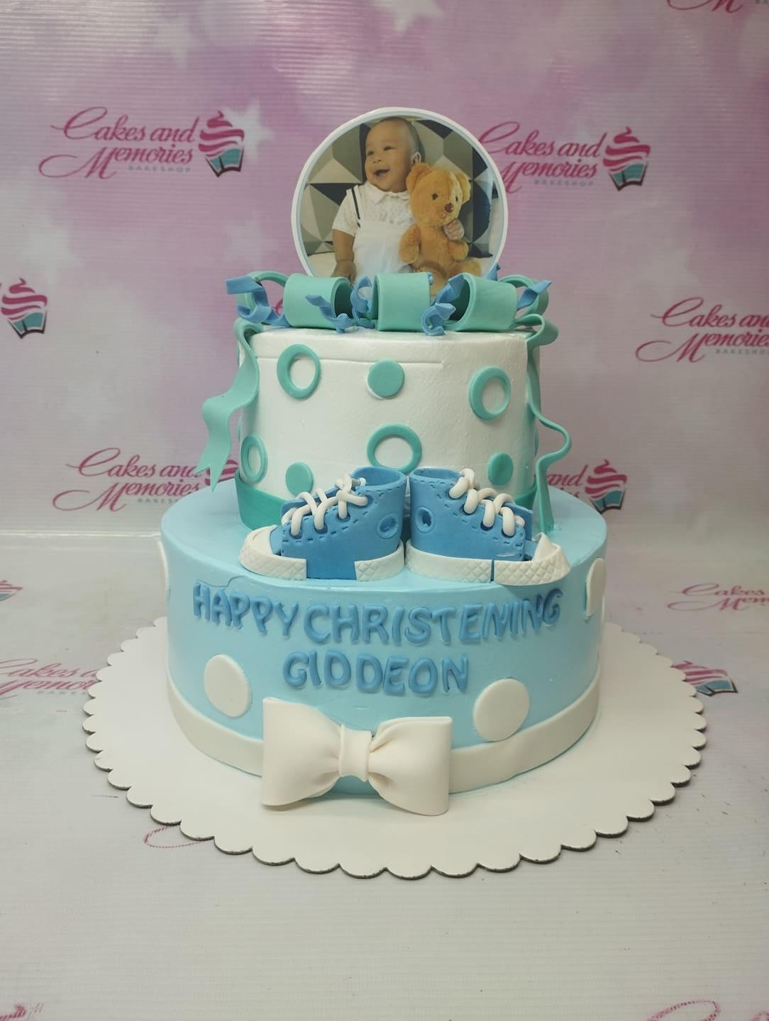 Blue and white 2-tier christening cake with fondant baby shoes, a bow, and a photo topper of a baby with a teddy bear.