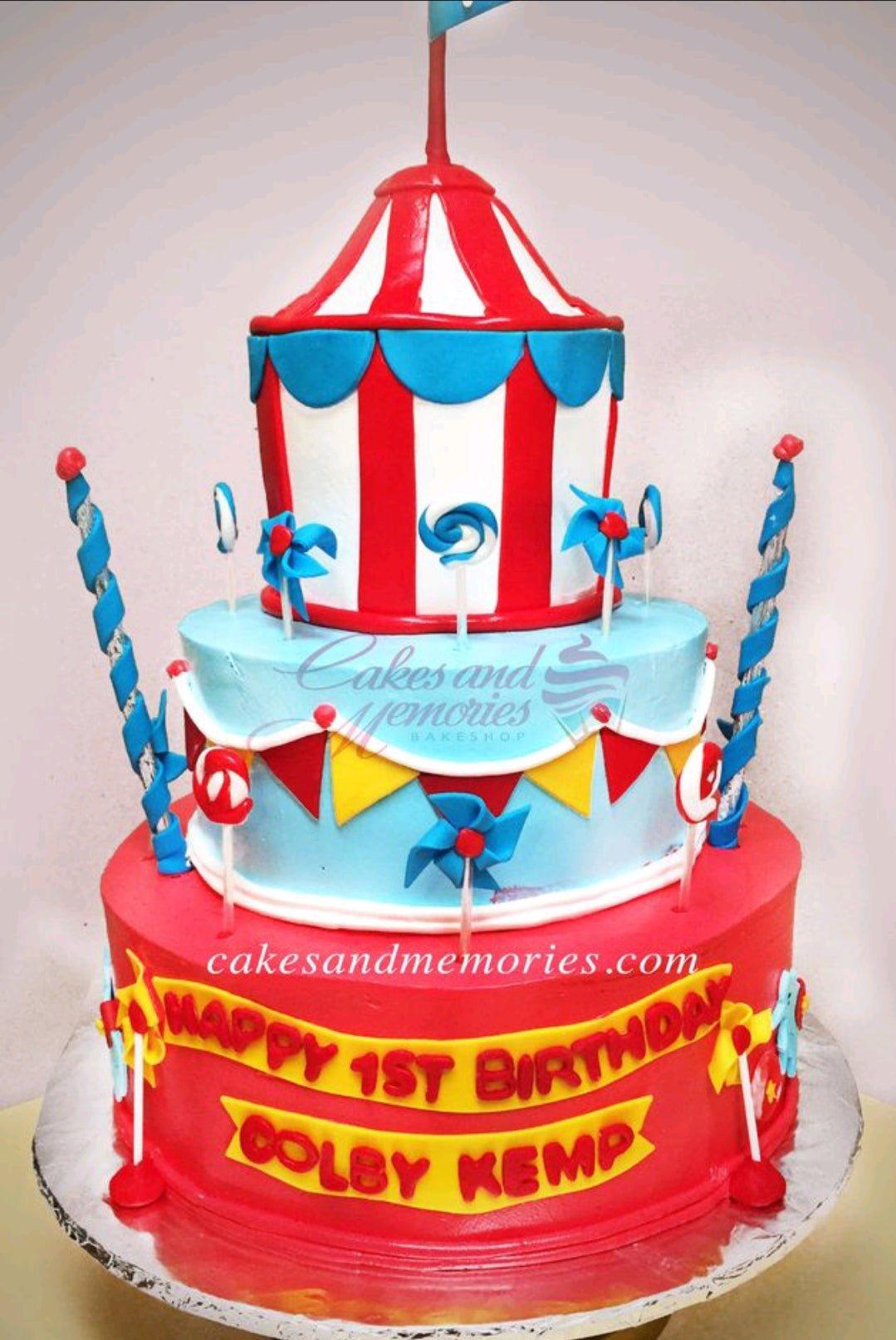 Three-tier circus-themed birthday cake with red and white stripes, blue bunting, and pinwheel toppers for a first birthday celebration.