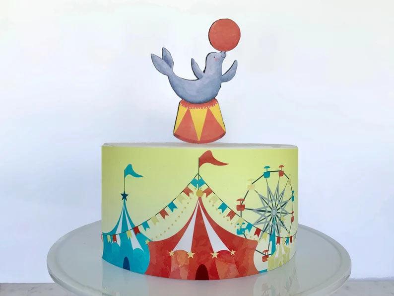 Circus themed birthday cake featuring a seal balancing a ball on its nose and a colorful carnival tent wrap design.