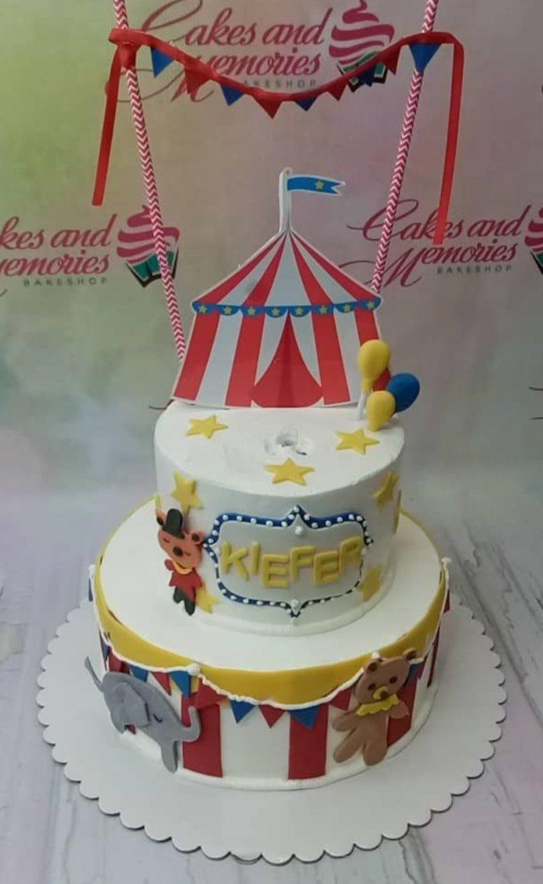 Two-tier circus themed birthday cake with a red and white striped tent topper, yellow stars, and fondant circus animals.