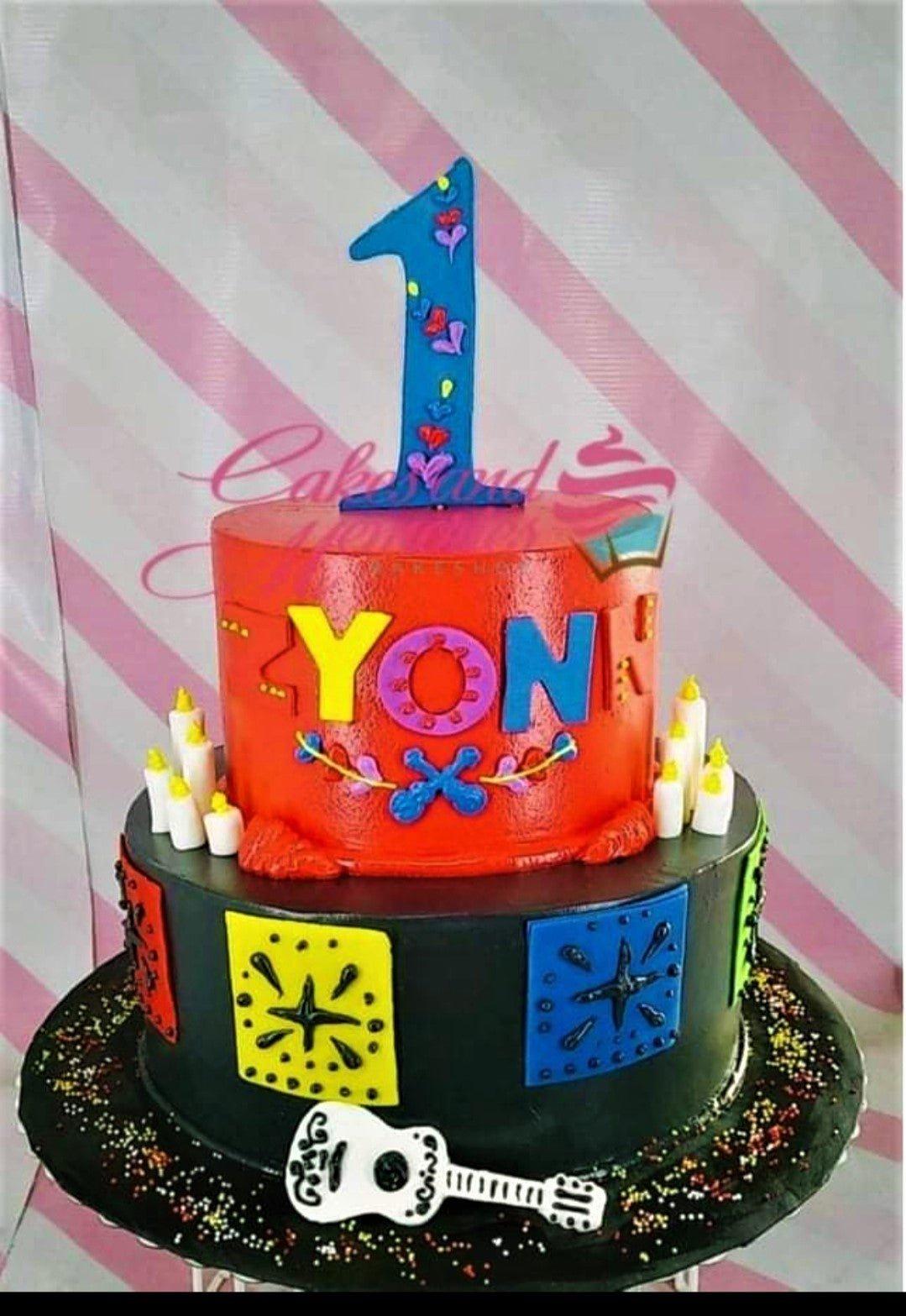 Two-tier Coco-themed birthday cake with red and black icing, featuring a blue number one topper and colorful papel picado designs.