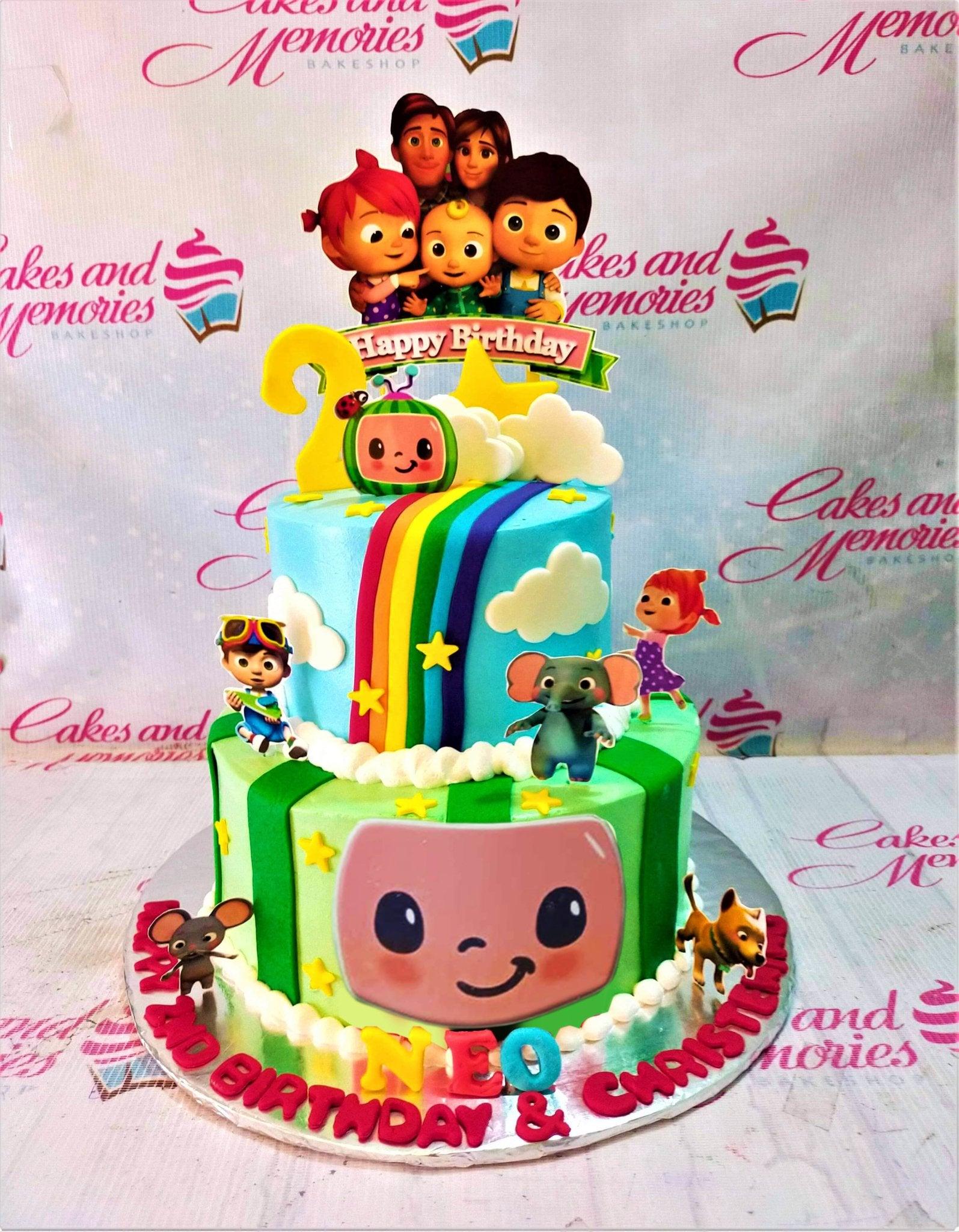 Two-tier Cocomelon themed birthday cake with blue and green icing, rainbow, clouds, and character printouts.