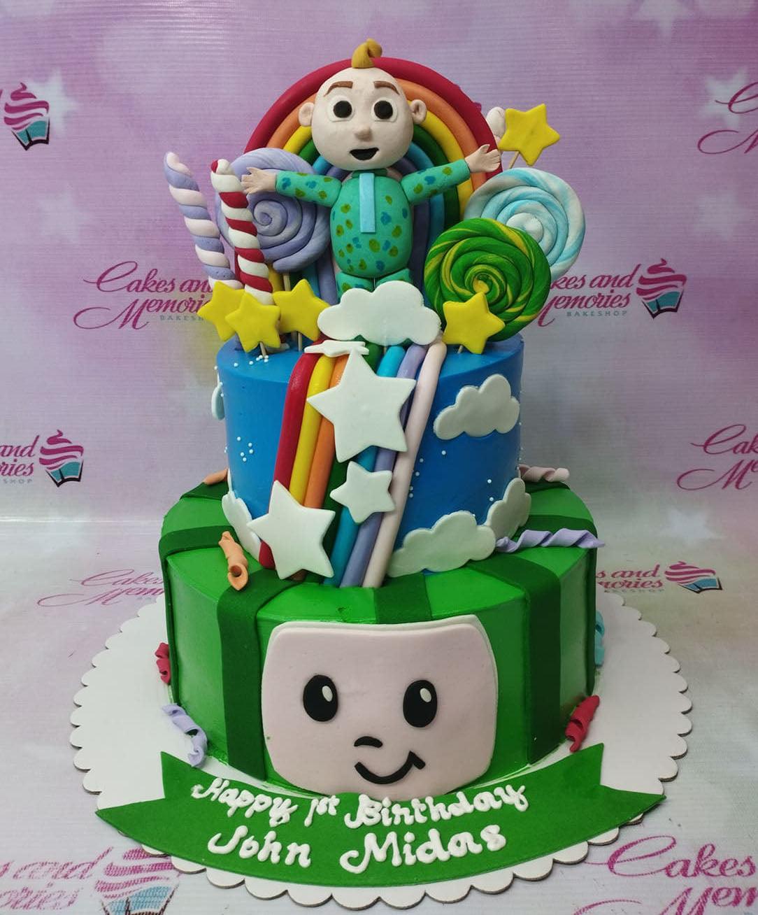 Two-tier Cocomelon themed birthday cake with a 3D JJ figure, rainbow, lollipops, and a green watermelon base tier.