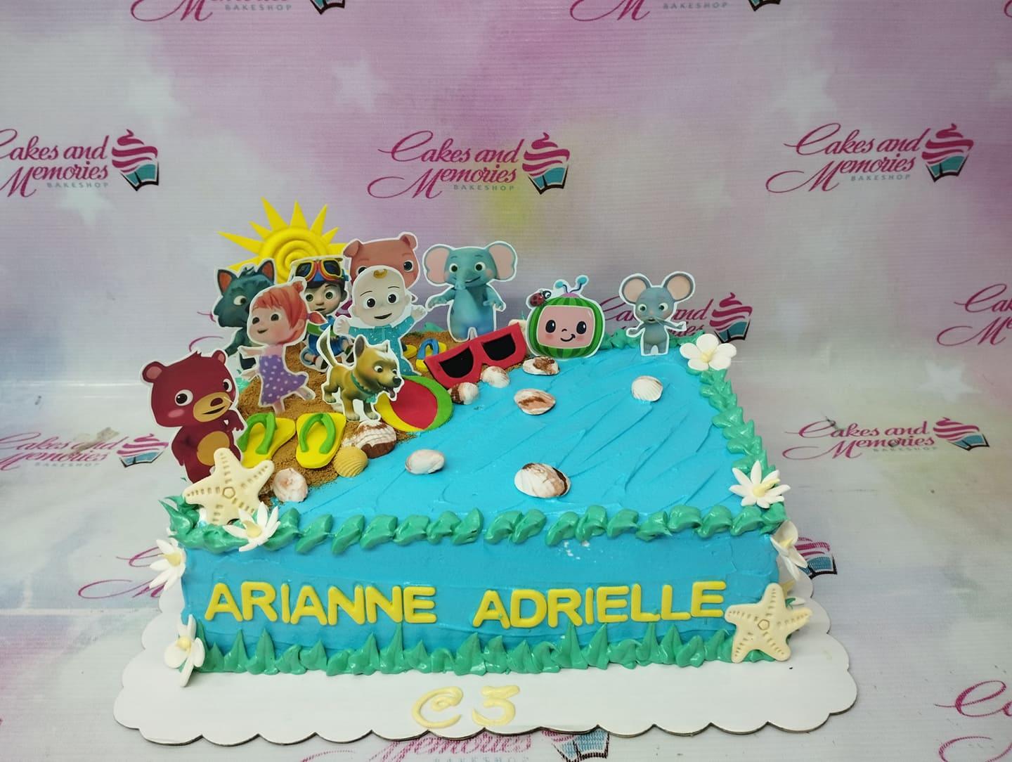 Cocomelon beach themed rectangle cake with blue icing waves, sand texture, and character printouts including JJ and friends.