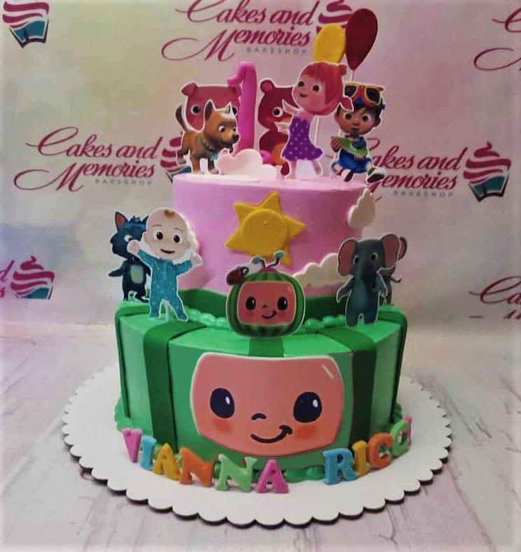 Two-tier Cocomelon themed birthday cake with pink top tier and green watermelon bottom tier featuring character printouts and fondant letters.