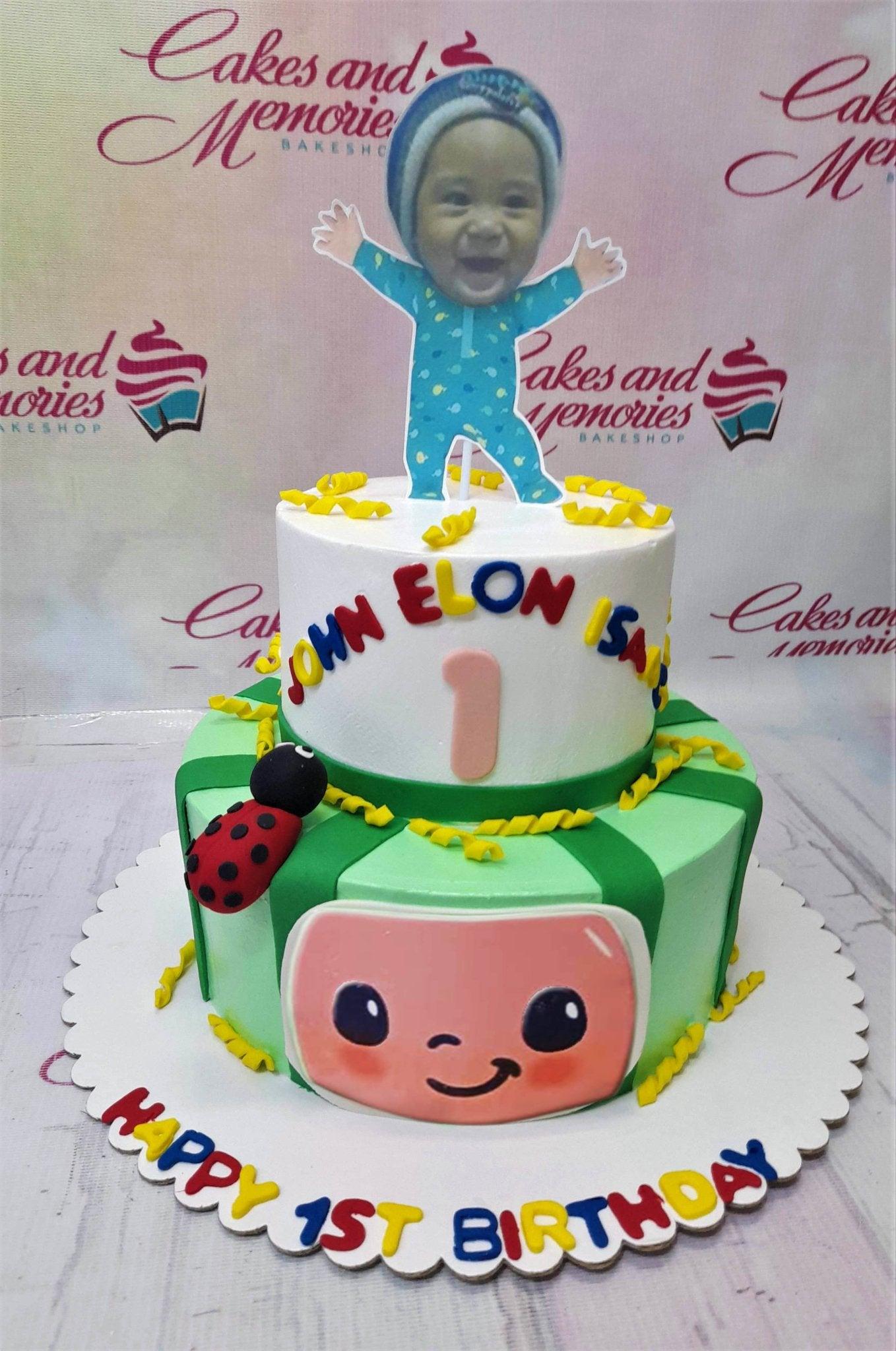 Two-tier Cocomelon themed birthday cake with a custom baby photo topper, ladybug, and colorful gumpaste letters.