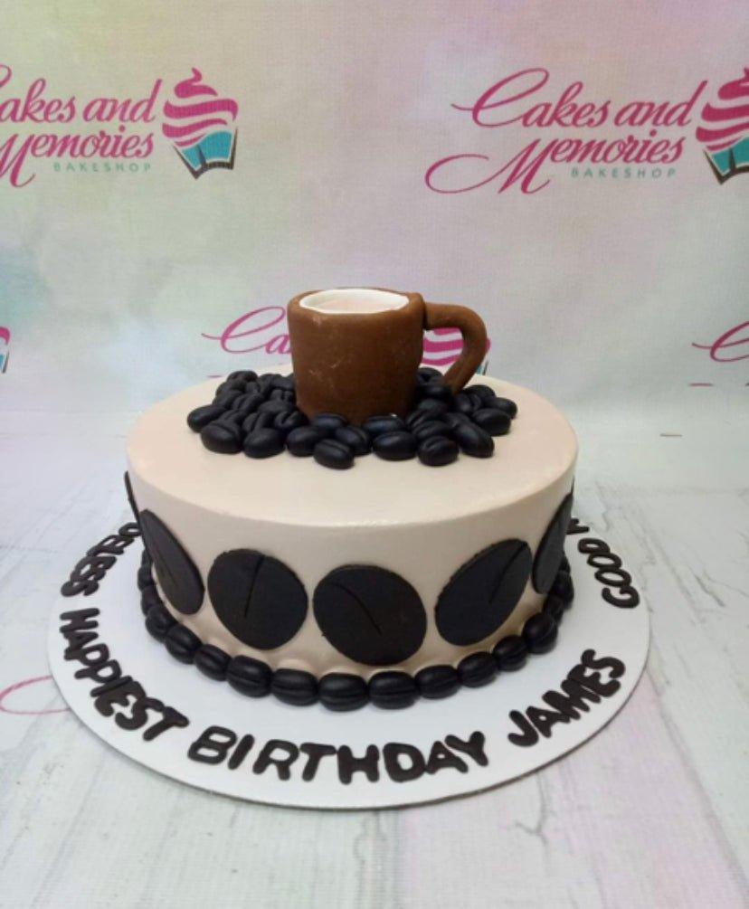 Coffee themed birthday cake with a brown fondant mug topper and black coffee bean decorations on a beige icing base.