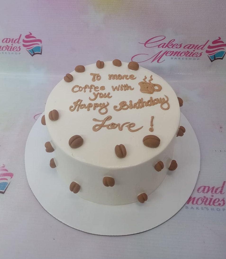 White minimalist birthday cake decorated with brown coffee bean fondant toppers and a coffee mug graphic.