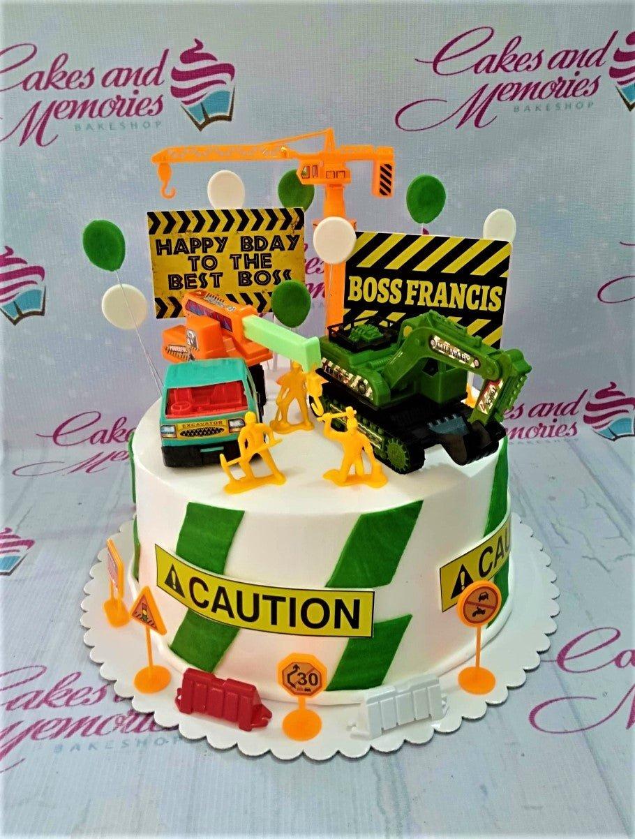 Construction themed birthday cake for Boss Francis with toy excavators, cranes, and caution signs on white icing with green stripes.