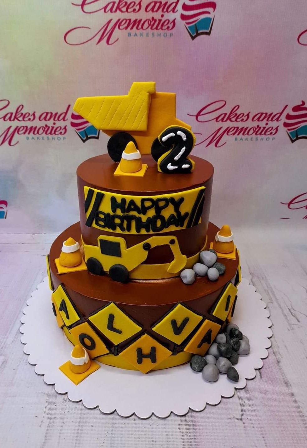 Two-tier brown construction themed birthday cake with yellow dump truck and excavator fondant toppers, traffic cones, and grey stones.