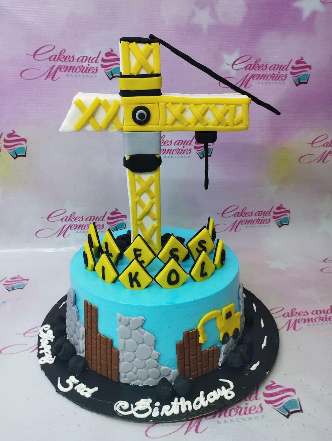 Blue construction themed birthday cake with a large yellow crane topper and gumpaste road and building details.