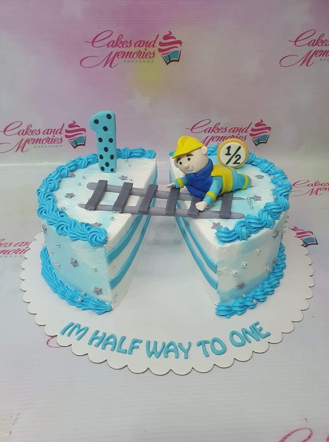 Half-cut light blue and white cake for a 6-month birthday featuring a construction worker figure on a ladder and a number 1 topper.