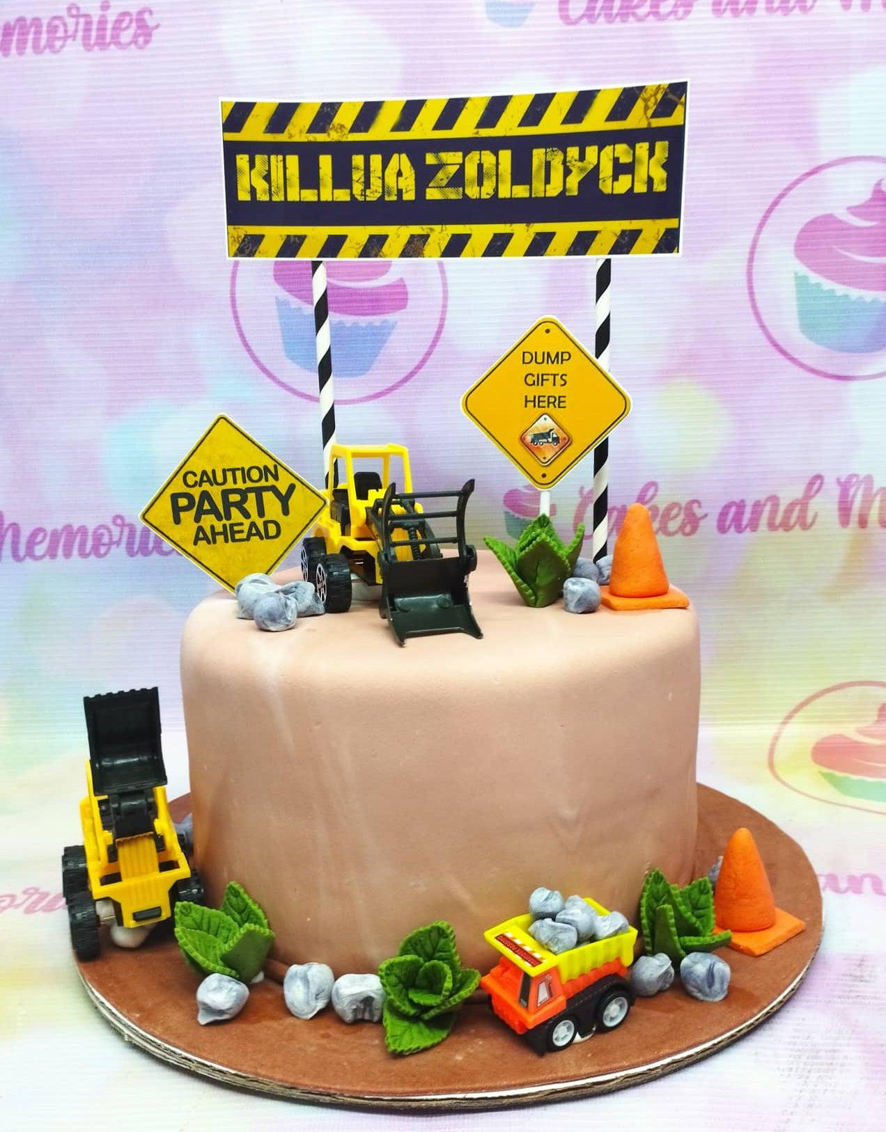 Tan fondant construction themed cake with toy trucks, orange cones, and a Killua Zoldyck construction sign topper.