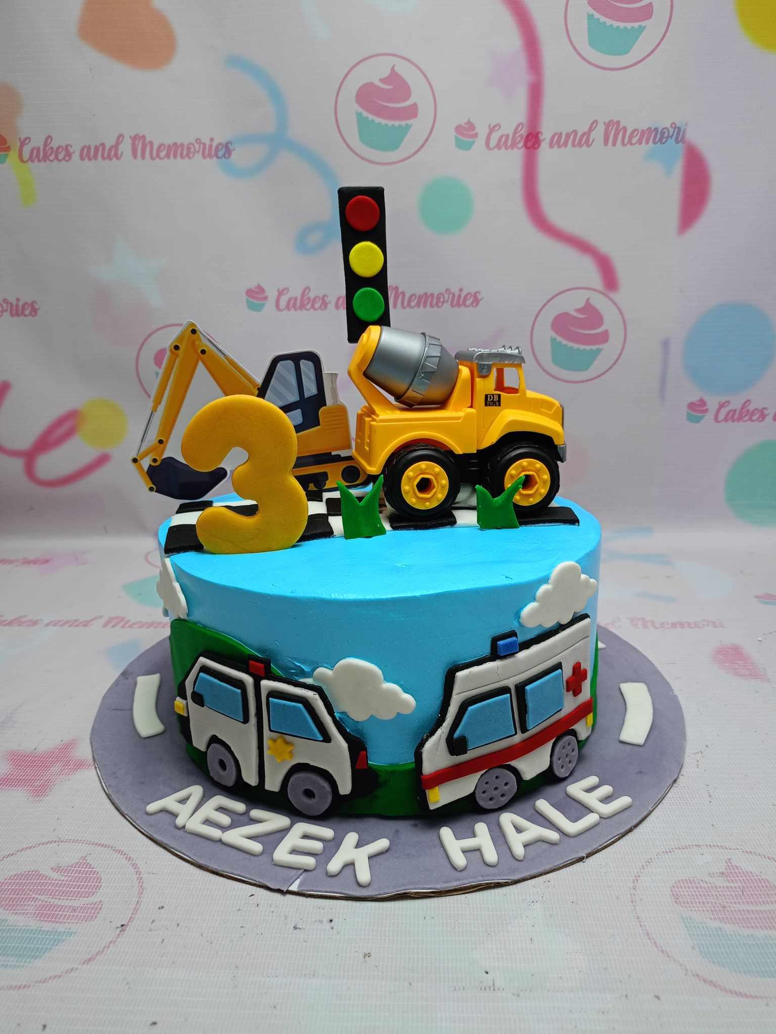 Blue construction and emergency vehicle themed birthday cake with toy excavator, cement mixer, and fondant ambulance and police car decorations.