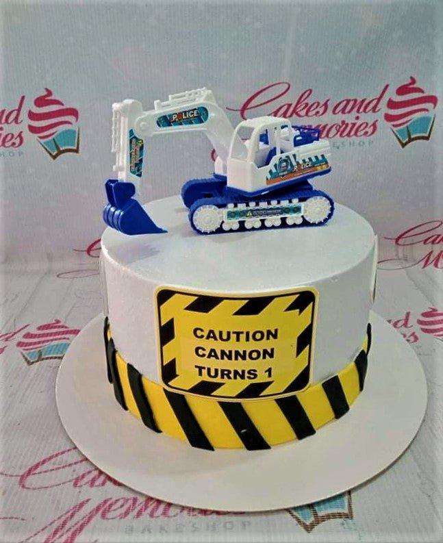 White construction themed birthday cake with a blue and white toy excavator on top and a yellow caution sign message.