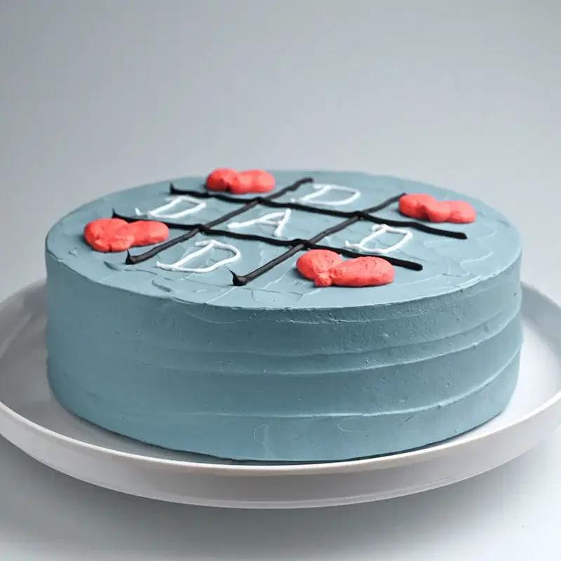 Blue bento cake with a black icing tic-tac-toe grid and red heart-shaped icing marks for a Father's Day celebration.