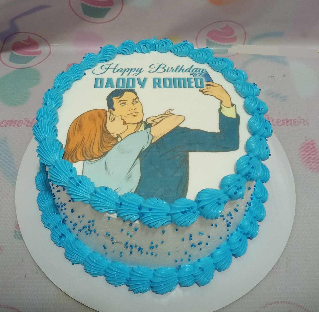 White round cake with blue shell borders and a comic-style printout of a couple taking a selfie for Daddy Romeo's birthday.