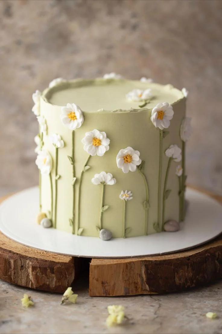 A minimalist light green cake decorated with delicate white piped icing daisies and green stems on a rustic wood base.