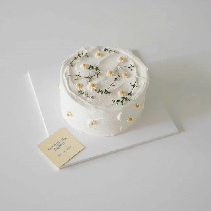 Minimalist white cake with delicate white and yellow daisy flowers and green herbal sprigs on a white square board.