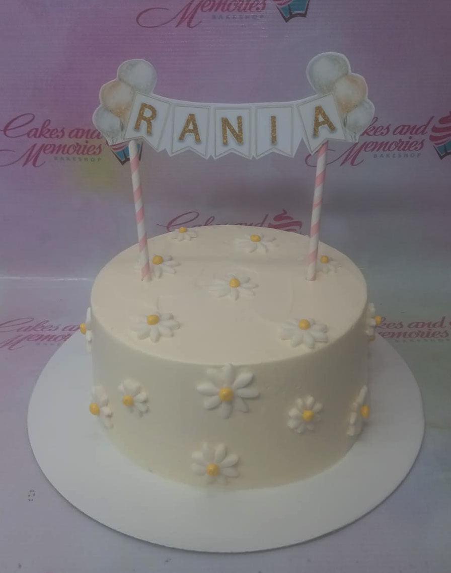 White minimalist cake decorated with small white and yellow icing daisies and a Rania name banner topper.