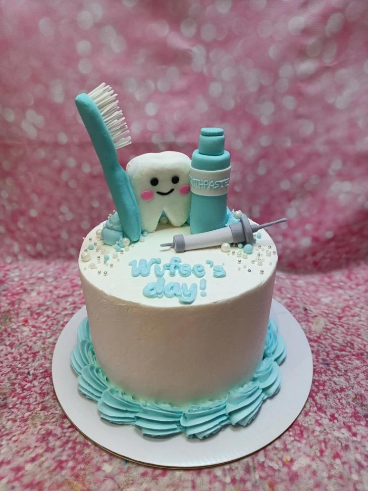 A white 1 Tier cake featuring fondant toothbrush, toothpaste, tooth, syringe, light blue icing swirls, piped border and white and silver sprinkles.