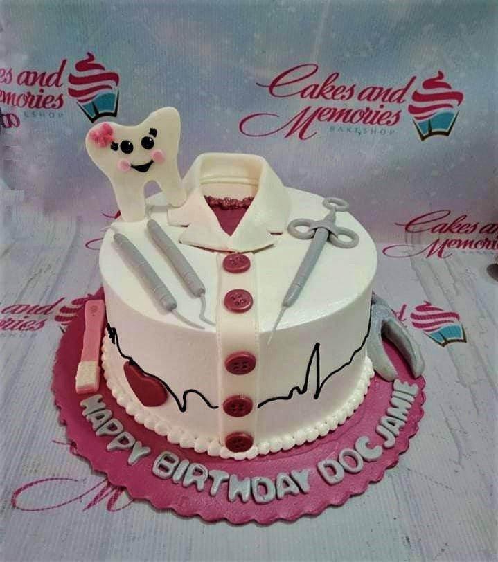White dentist themed birthday cake with a fondant tooth character, medical tools, and a doctor's coat design on a pink base board.