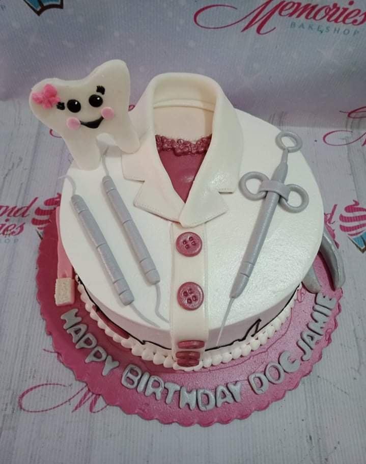 White fondant dentist themed cake with a 3D tooth character, dental tools, and a doctor's coat design on a pink base board.