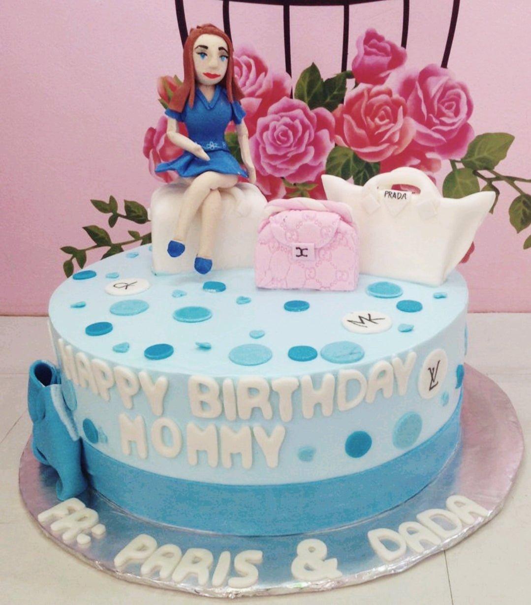 Light blue fondant cake with a 3D fondant woman figure sitting on luggage, pink and white designer bags, and blue polka dots.