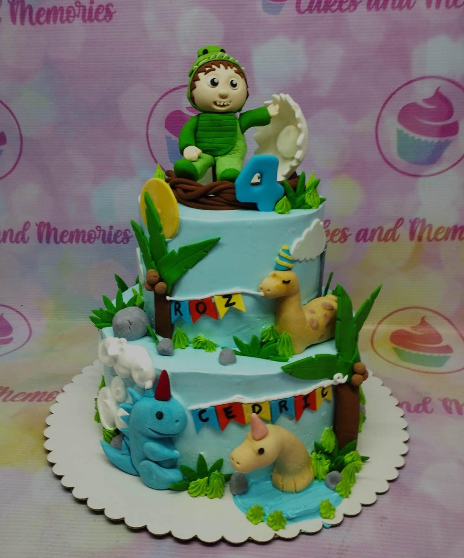 Two-tier light blue dinosaur themed birthday cake with fondant figures of a boy in a dinosaur suit, dinosaurs, and palm trees.