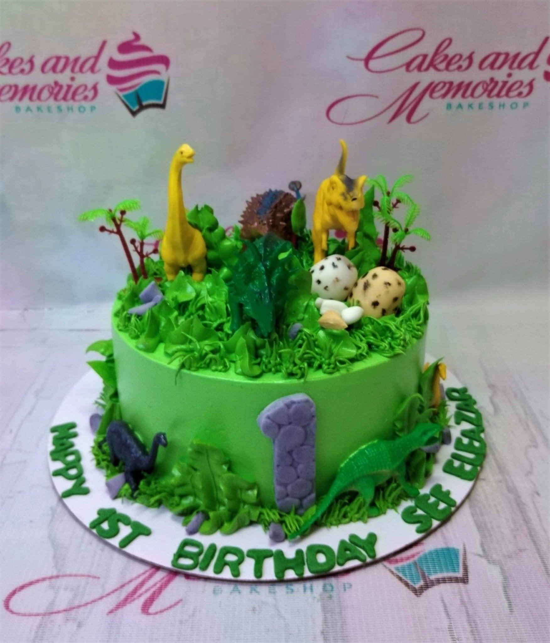Green dinosaur themed birthday cake with 3D fondant dinosaurs, eggs, and trees on a grass-textured icing base.