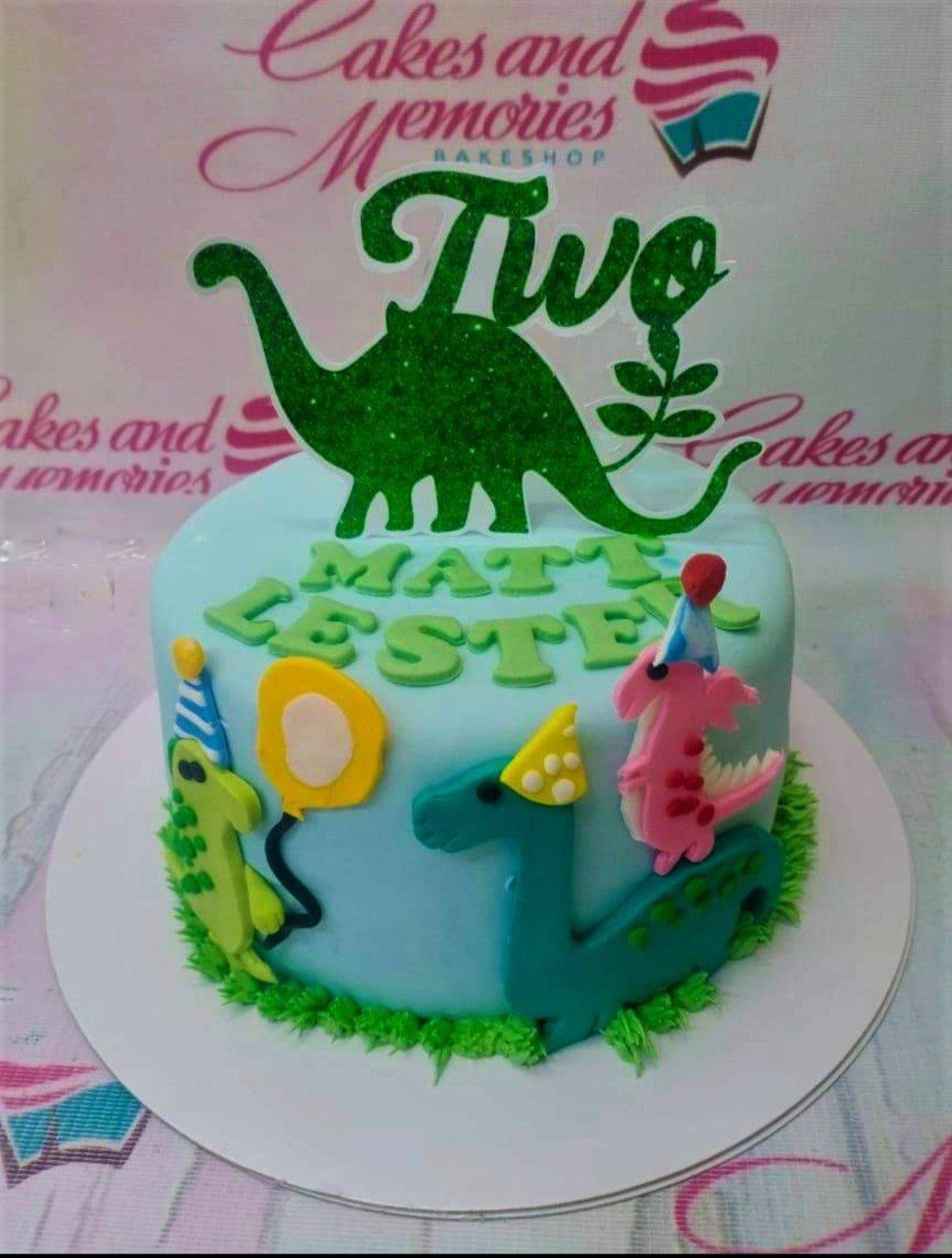 Light blue dinosaur themed birthday cake with colorful fondant dinosaur figures and a green glittery 'Two' topper.