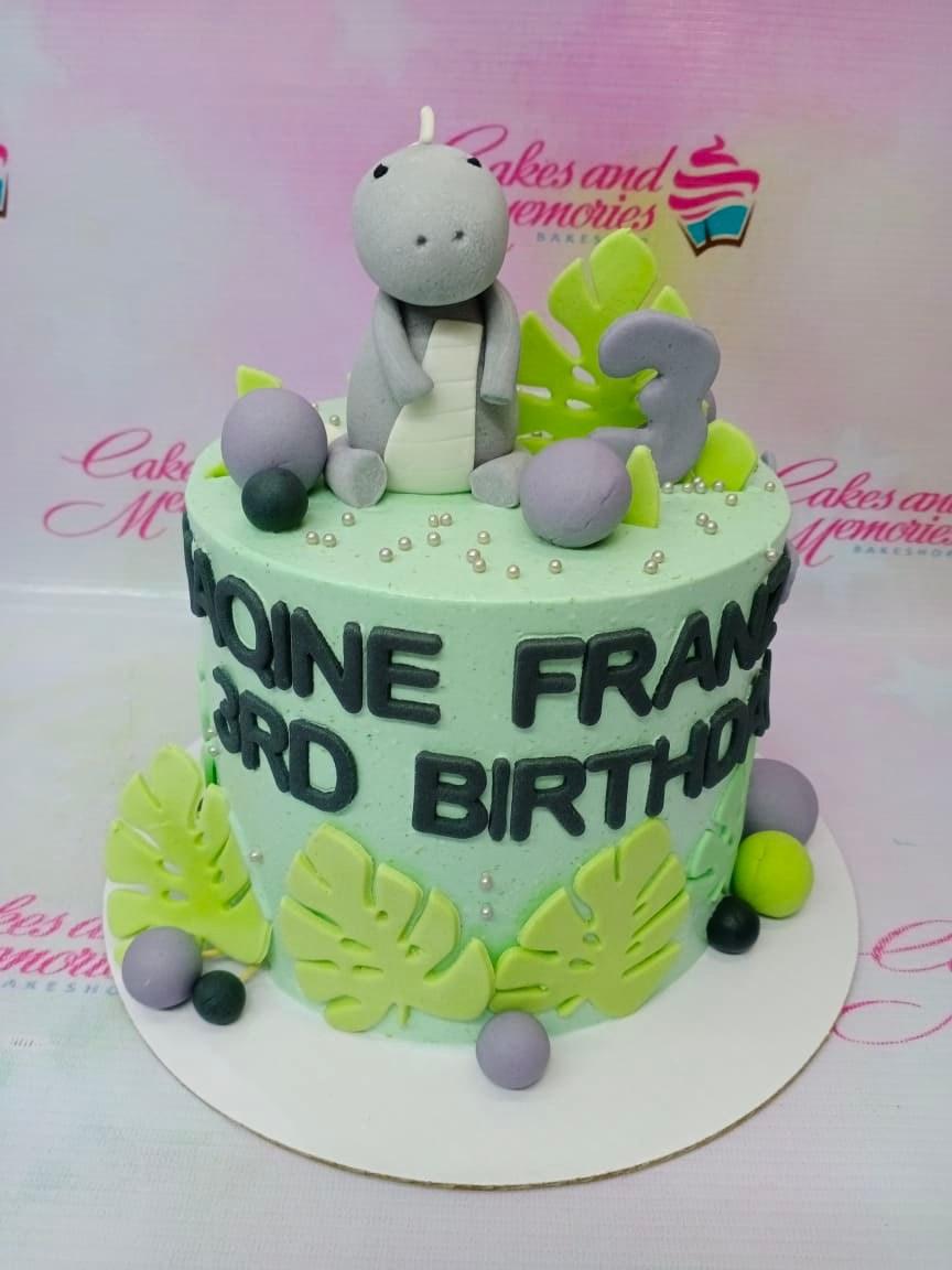Light green 1-tier dinosaur birthday cake with a grey fondant dinosaur topper, green tropical leaves, and grey fondant balls.