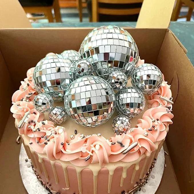 Pink disco ball cake with peach icing swirls, white chocolate drip, and silver sprinkles in a cardboard box.