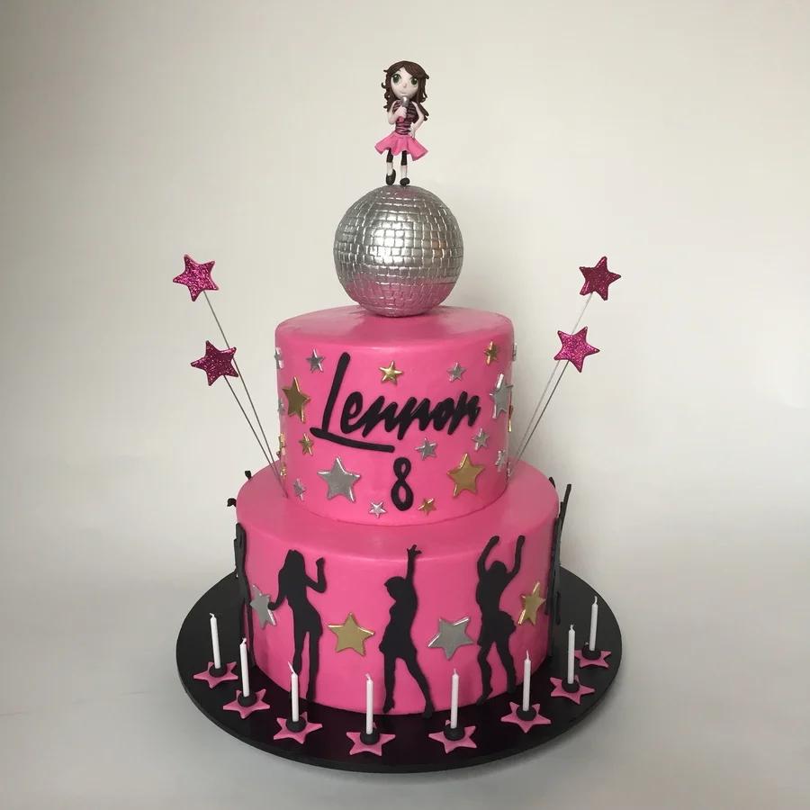 Pink 2-tier disco themed birthday cake with a silver disco ball, a girl figurine, and black dancer silhouettes.