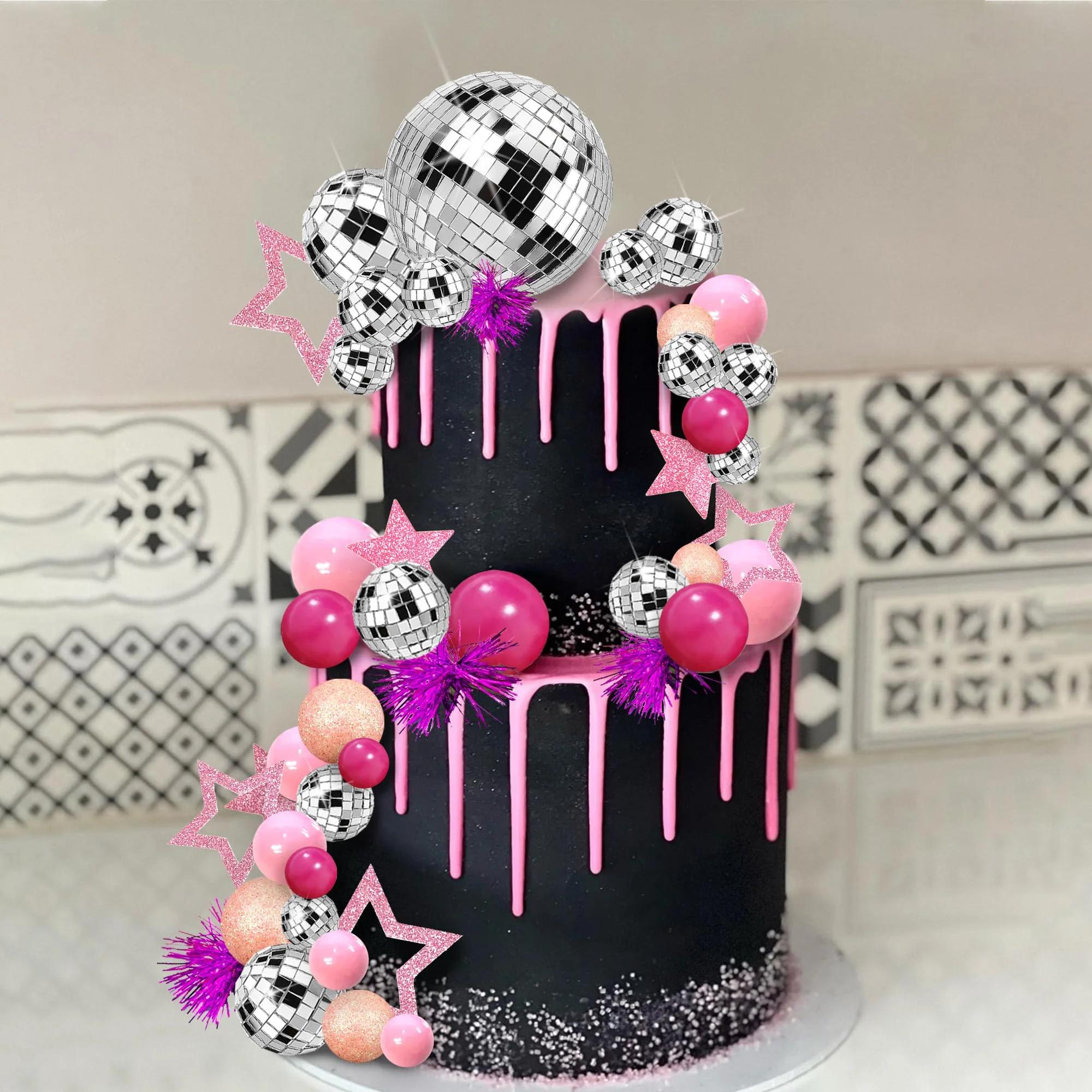 Two-tier black cake with pink drips, disco balls, pink stars, and purple tinsel accents.